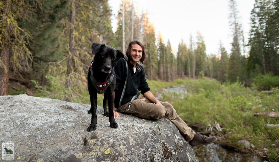 Working dog in custom harness and handler sit on rock during break in the woods.
