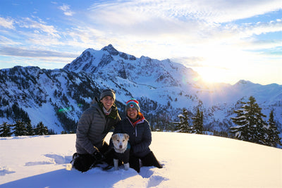 A woman and man sit with their dog on a snowy mountain. 