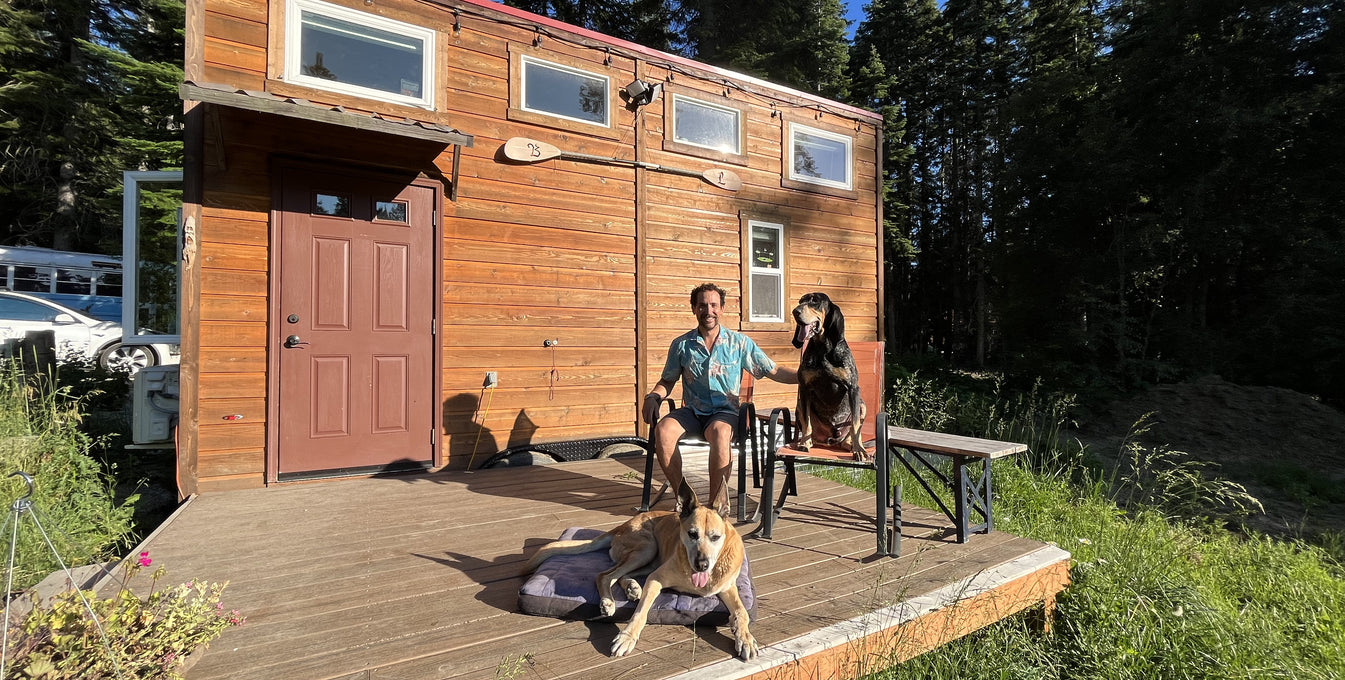 A man and his two dogs sit on the front porch of their tiny house.
