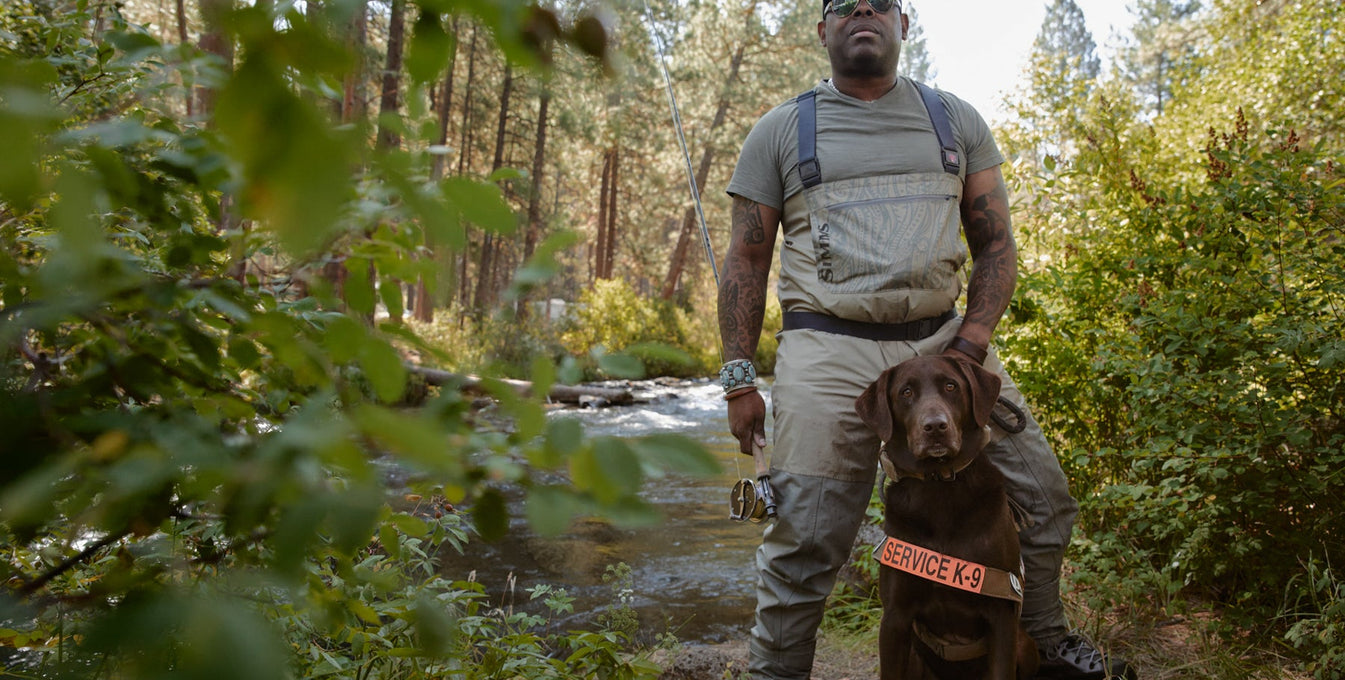 Ruffwear ambassador Chad stands in fishing waders with service dog Axe next to river.
