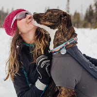Dog in cloud chaser jacket licks human's face on snowy day outside.