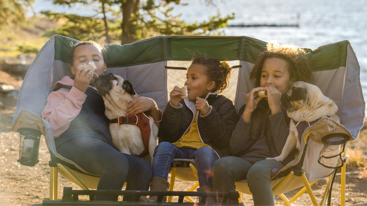 Three girls sit on a big camp chair with two pugs eating s'mores next to a campfire along a lake while camping,.