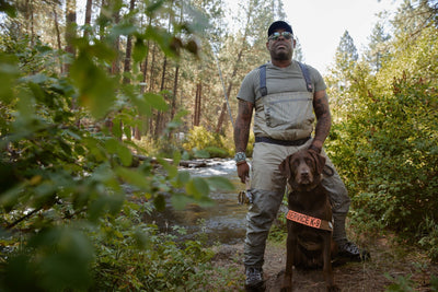 Chad in fly fishing gear and dog axe in service dog vest pose by the river.