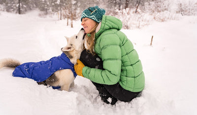 Becca in puffy jacket in snow gets kisses from Tala the husky in Quinzee insulated jacket.