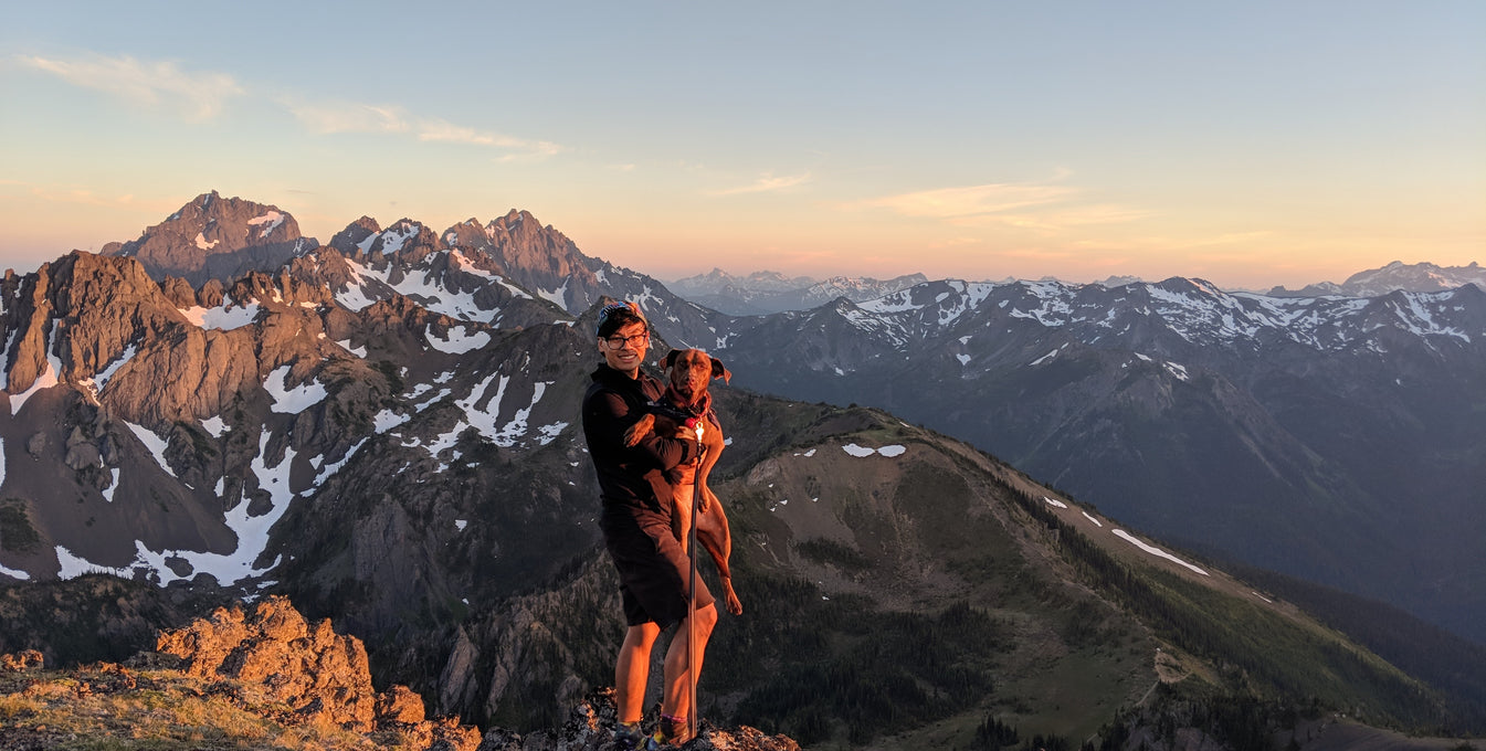 A man holds his dog while standing on the top of a mountain trail. 