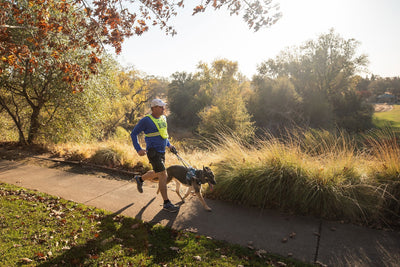 Blind runner and dog in Unifly run along a paved trail with trees.