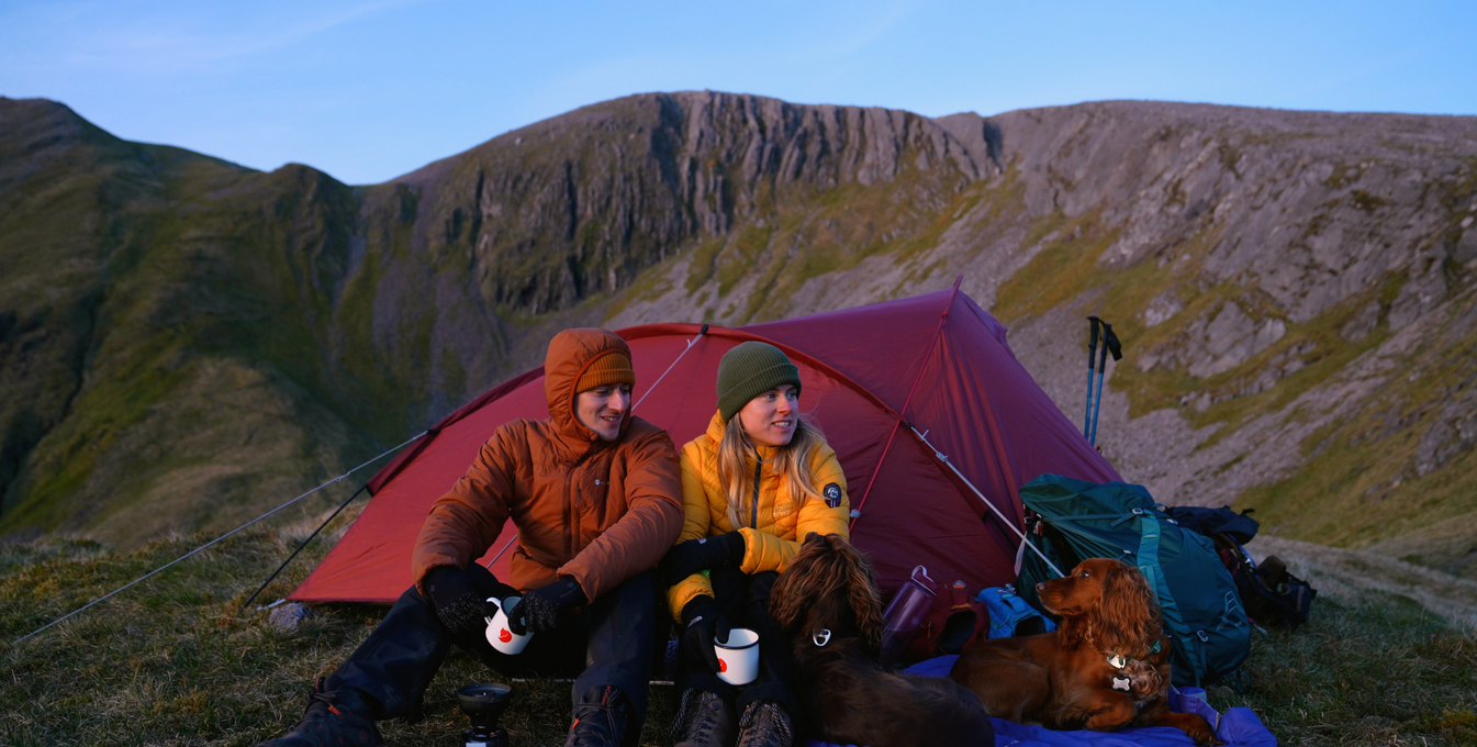 Man and woman and two spaniel dogs sitting in front of a tent in the Scottish Highlands
