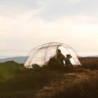 Dog and human in tent at golden hour in grassy field.