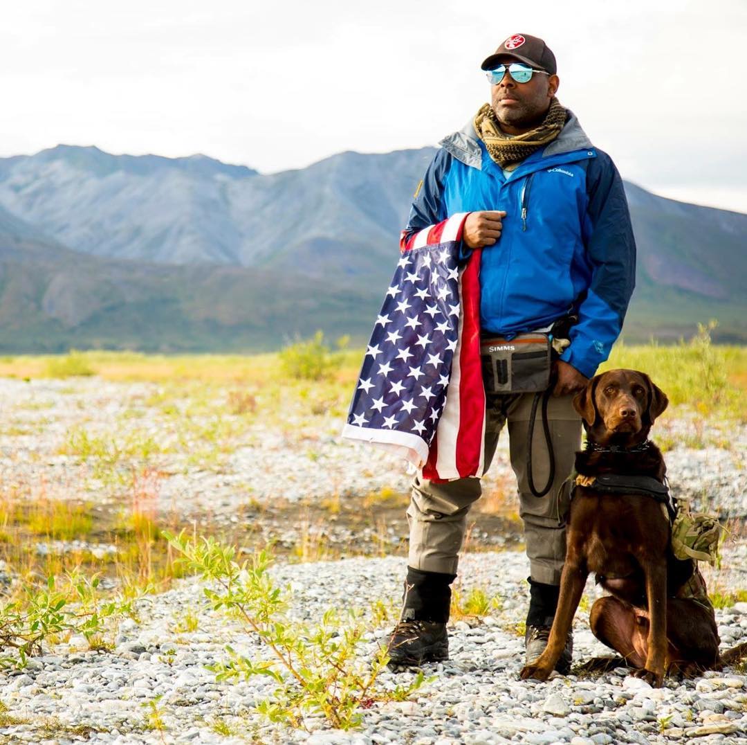 Chad holds an american flag and stands next to his service dog axe.