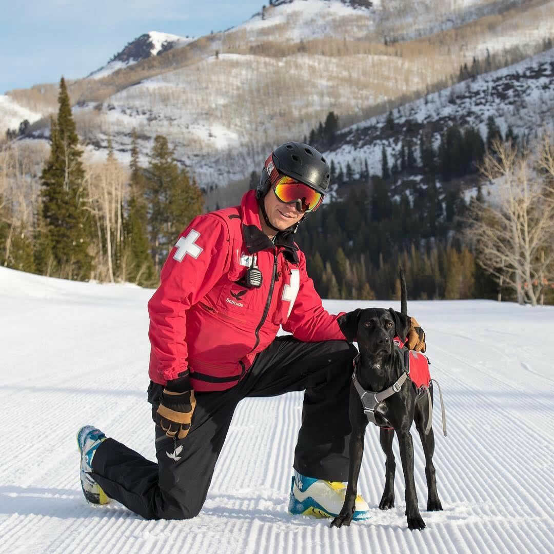 Avalanche dog and patroller kneel on corduroy groomed run in the mountains.