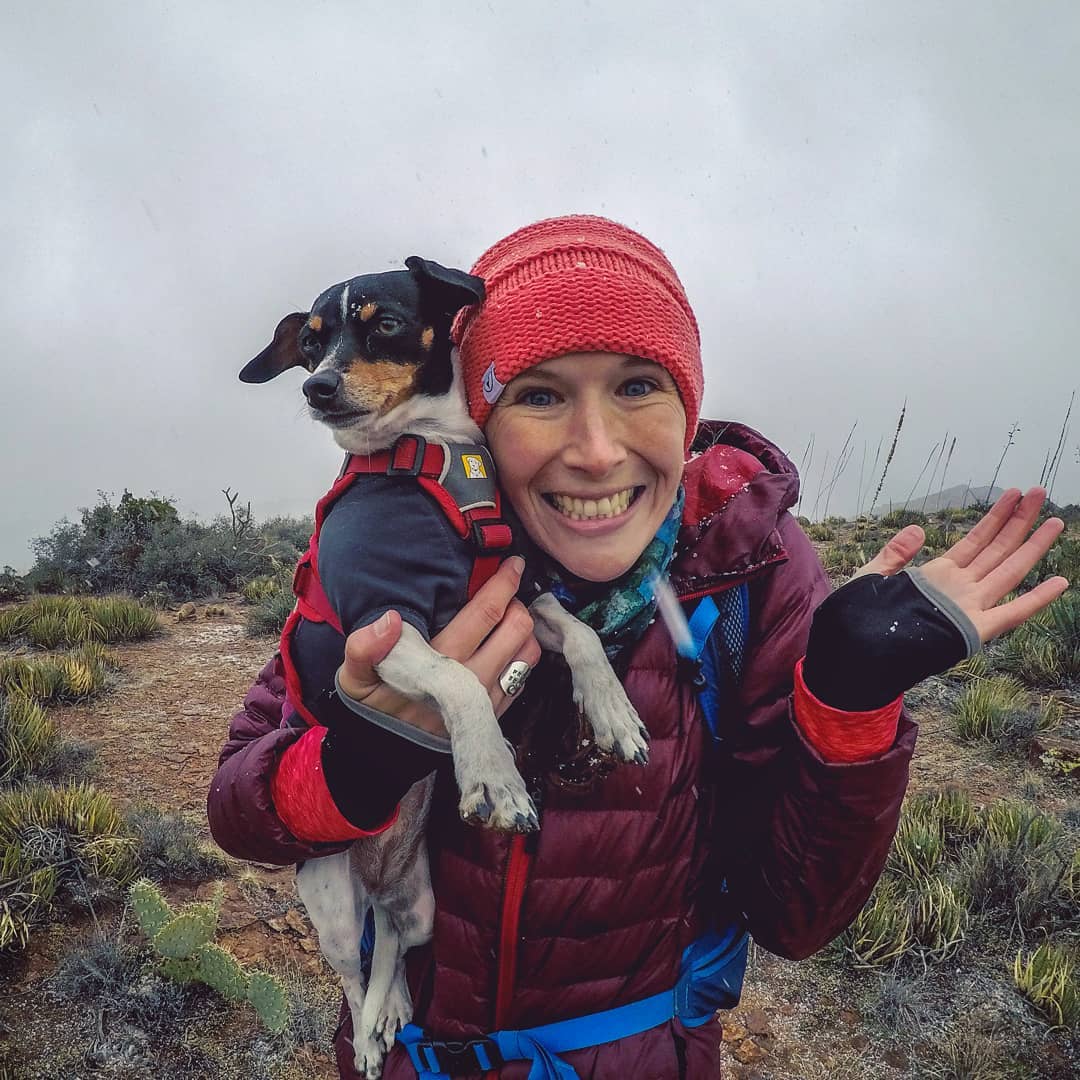 Katie holds her little dog up next to her face smiling with excitement on a hike.