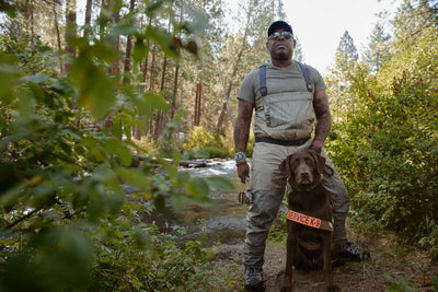 Ruffwear ambassador Chad stands in fishing waders with service dog Axe next to river.