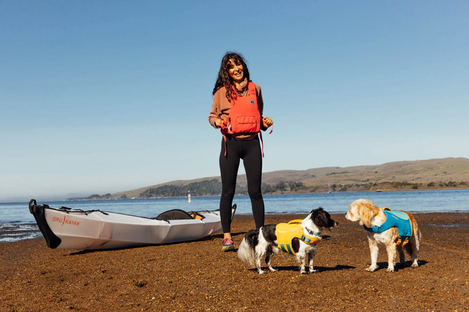 Noel stands next to her two dogs and a kayak on a beach.